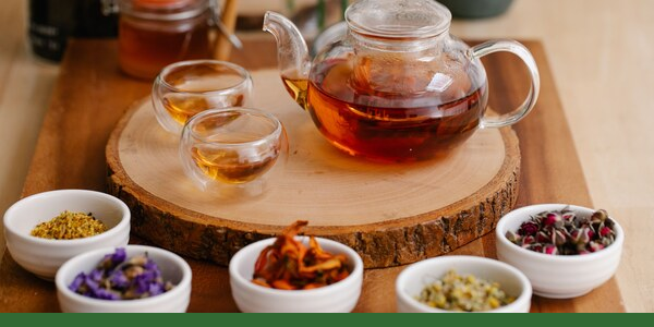 Glass teapot and cups of herbal tea with bowls of dried botanicals and a Gaia Garden green line along the bottom